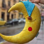 Cojín infantil con forma de luna en color amarillo. La luna lleva un gorrito de dormir para invitar al sueño de los niños.